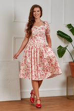 Load image into Gallery viewer, Woman wearing a floral dress standing in a room with a plant and decorative items.