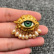 Load image into Gallery viewer, Gold eye-shaped pendant with green gemstone and pearl decorations held between fingers on a textured gray background.