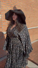 Load image into Gallery viewer, Woman wearing a black hat and patterned dress standing against a brick wall.