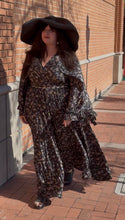 Load image into Gallery viewer, Woman in a patterned dress and large black hat standing on a brick sidewalk.