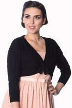 Load image into Gallery viewer, A woman modeling a black knit wrap cardigan with a front twist detail, worn over a light pink dress against a plain white background.