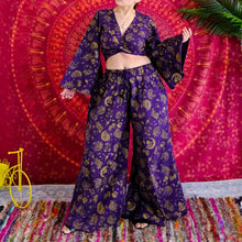 Load image into Gallery viewer, A woman models a purple and gold printed two-piece outfit consisting of a wrap-style crop top with wide bell sleeves and matching high-waisted wide-leg pants, posed in front of a red and pink bohemian mandala tapestry.