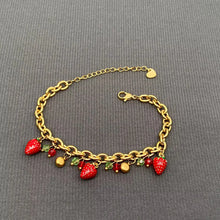 Load image into Gallery viewer, A gold-tone chain bracelet with dangling strawberry charms, red and green enamel details, and small gold beads, displayed on a dark gray textured fabric background.
