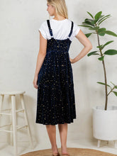 Load image into Gallery viewer, A woman stands with her back to the camera wearing a navy blue velvet pinafore dress with gold star prints over a white t-shirt. The setting is a bright, modern room with a white stool, a potted plant, and a textured white wall.
