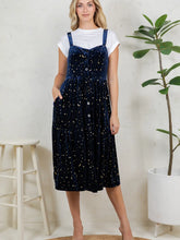 Load image into Gallery viewer, A woman models a navy blue velvet pinafore dress with a gold celestial print, layered over a white t-shirt. She is standing in a brightly lit room with a white stool on the left and a potted plant on the right.
