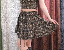 Load image into Gallery viewer, Person wearing a black and gold patterned outfit with a flowing skirt against a textured fabric background.
