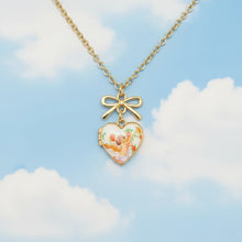 Load image into Gallery viewer, Gold chain necklace with a heart-shaped locket pendant featuring a small cherub illustration and a bow charm, displayed on a blue  sky background
