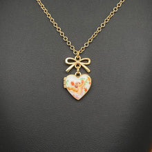 Load image into Gallery viewer, Gold chain necklace with a heart-shaped locket pendant featuring a small cherub illustration and a bow charm, displayed on a dark textured surface.
