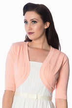 Load image into Gallery viewer, A woman wearing a light peach pink knit shrug cardigan over a white dress, photographed against a plain white background.
