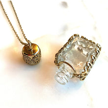 Load image into Gallery viewer, A gold-toned necklace with two pendant charms: one cylindrical filigree pendant and one small clear glass bottle-shaped pendant with a gold-toned textured border, displayed on a white marble surface.
