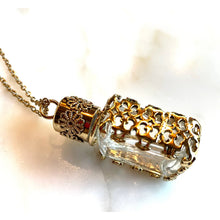 Load image into Gallery viewer, A gold-toned filigree pendant necklace featuring a small clear glass vial, shown on a white marble surface.
