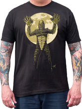 Load image into Gallery viewer, A man wearing a black graphic t-shirt featuring a green creature standing in front of a large yellow moon, with tattooed arms visible.
