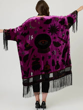 Load image into Gallery viewer, A woman viewed from behind wearing a bright magenta-purple velvet poncho with black occult-themed prints and long fringe trim, posed against a light gray studio background.
