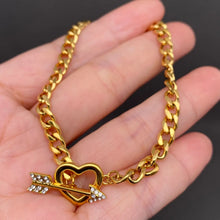Load image into Gallery viewer, A gold-tone Cuban link chain bracelet with a heart-shaped charm and arrow detail, held in a person's hand against a dark gray background.
