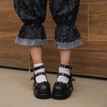 Load image into Gallery viewer, Black floral-patterned pants with lace cuffs worn by a person standing on a wooden floor.
