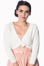 Load image into Gallery viewer, A woman wearing a white knit cardigan with a bow detail at the neckline over a light pink dress, photographed against a plain white background.
