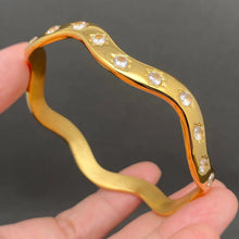 Load image into Gallery viewer, A gold bangle bracelet with a wavy, undulating line and small clear gemstones set along the top, held in a person's hand against a dark gray background.
